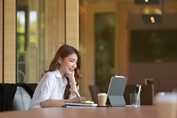 Photo of a young businesswoman keeping a hand on her chin while taking notes at the wooden meeting table surrounded by a digital tablet and office equipment in the comfortable meeting room.