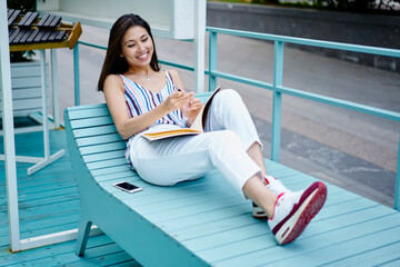 Joyful female student with education sketchbook laughing during learning time in city, funny Asian blogger with textbook resting at blue garden chair and smiling while creating publication ideas