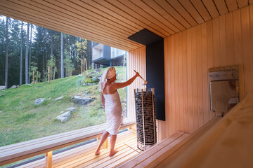 Relaxing Woman wrapped white towel in Hot Finnish sauna with huge wide window with green forest...
