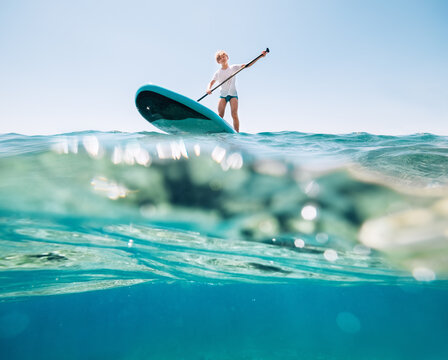 Under the water view angle to the smiling blonde teenager, boy rowing stand up paddleboard. Active family summer vacation time near the sea concept image.