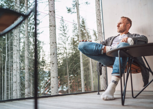 Portrait of Middle-aged man dressed open cardigan, jeans, and warm socks sitting on forest house balcony and enjoying the fresh air. Everyday lifestyle and enjoying the Nature concept image.