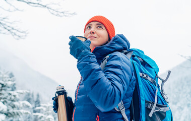 Middle-aged woman smiling and drinking a hot drink from thermos flask dressed warm down jacket...