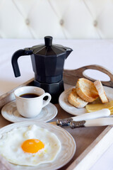 A breakfast tray with a fried egg, a cup of black coffee and a coffee pot is on a white bed.  Theme breakfast in bed. Vertical image.