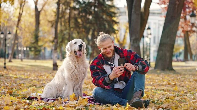 Funny content. Young positive bearded guy taking selfie with his dog on smartphone during walk in autumn park