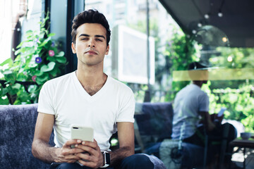 Portrait of handsome Caucaisan blogger with digital mobile device looking at camera during weekend pastime at street cafeteria terrace, millennial generation Z with modern smartphone posing