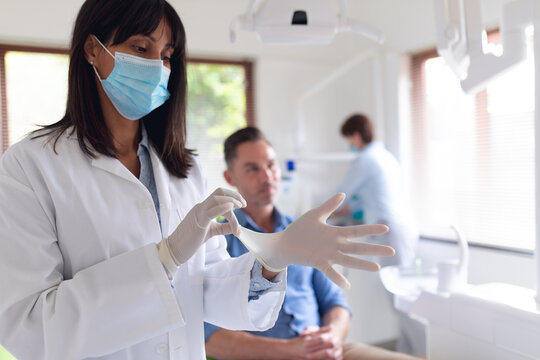 Biracial female dentist wearing medical gloves and male patient waiting at modern dental clinic