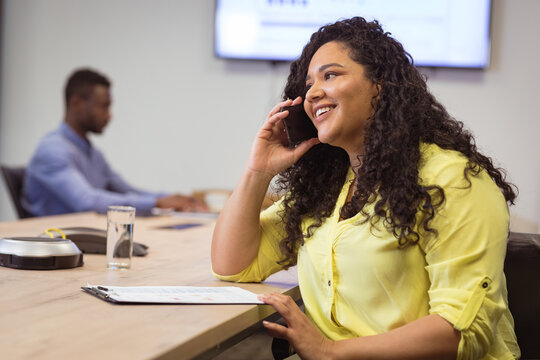 Biracial smiling businesswoman talking on smartphone and sitting at table in modern office