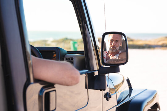 Portrait of smiling caucasian man in car reflected in side mirror on sunny day at seaside