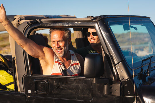 Happy caucasian gay male couple waving, sitting in car on sunny day at seaside