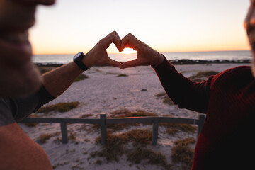 Happy caucasian gay male couple forming heart shape with hands on beach at sunset