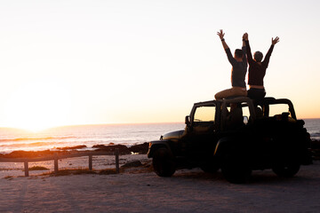 Caucasian gay male couple sitting on car roof raising arms and holding hands at sunset by the sea