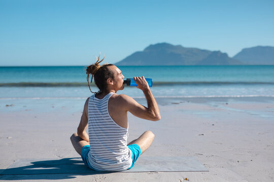 Rear view of man drinking water from bottle while doing yoga at beach on sunny day
