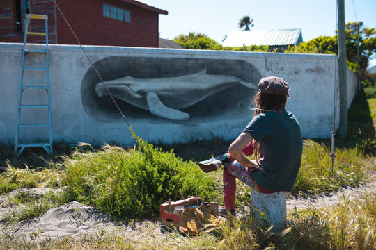 Male artist sitting while admiring his beautiful whale mural on wall during sunny day - Powered by Adobe