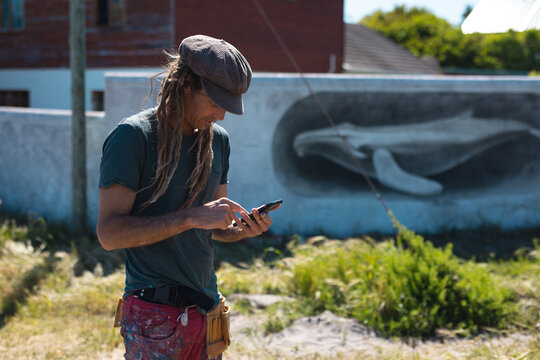 Male mural artist using smart phone while standing near whale painting on wall during sunny day