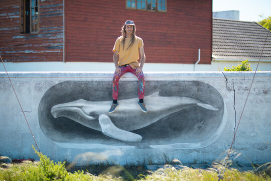 Portrait of male artist sitting on wall with whale mural painting against house