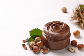 Glass jar with tasty chocolate spread, hazelnuts and green leaves on light background