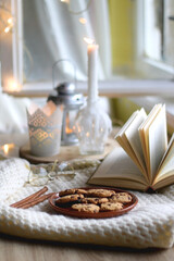Plate of chocolate chip cookies, cinnamon sticks, soft blanket, open book and lit candles. Hygge at home. Selective focus.