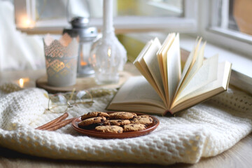 Plate of chocolate chip cookies, cinnamon sticks, soft blanket, open book and lit candles. Hygge at home. Selective focus.