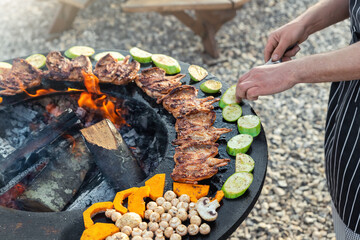 Close-up detail view of chef flipping by tongs tasty crispy marinated quails and vegetables grilled at round steel iron firepit hearth table surface brazier with burning firewoods. Barbecue yard home - obrazy, fototapety, plakaty