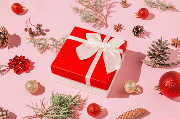 Creative composition made of red present box with white bow on pastel pink background with juniper branch, baubles and pine cones. Christmas gift. Celebration and party concept.