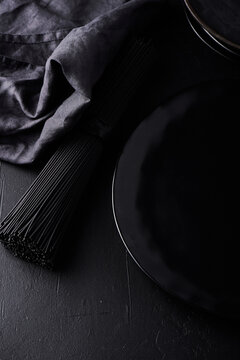 Dark food background with empty black stone board, ceramic plate, raw nero di seppia spaghetti and textile napkin. Free space for text or product. Top view, flat lay. Copy space