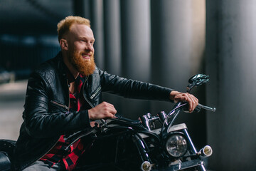 Fototapeta na wymiar Portrait of smiling biker. Young bearded man sitting astride motorbike. Medium shot.