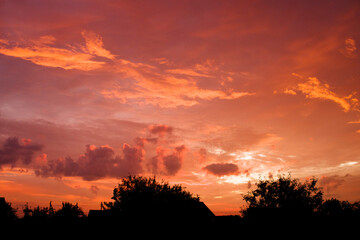 Red and orange cloudy sky before sunrise against a black silhos of rustic roofs and trees. August Landscape with Dawn Sky