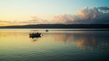 Obraz premium Couché de soleil sur un petit bateau sur un lac, les nuages se reflètent sur l'eau, de couleur orange et bleu. Une colline en face.