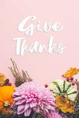 Give thanks text on pumpkins, dahlias flowers, leaves, heather on pink background flat lay. Happy thanksgiving Seasonal greeting card, handwritten sign. Thanksgiving greetings