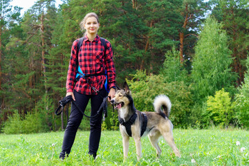 Naklejka na ściany i meble A young girl in a red shirt with her dog stands against the background of the forest. Walking with a pet. Copy space