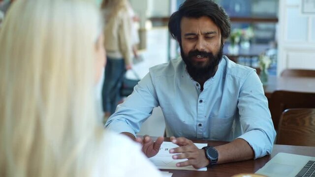 Close-up back view of female applicant during job interview talking to young male employers. Businesswoman sitting at desk interviewing bearded smiling male candidate in cozy coffee shop, slow motion.
