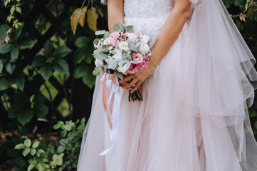 A bride in a long white and pink dress holds a wedding bouquet of flowers with a ribbon.