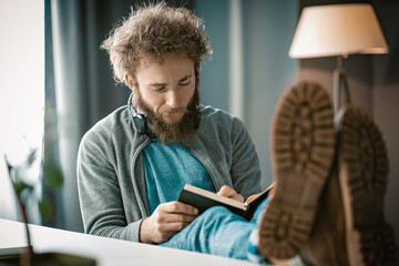 Curly-haired Guy Writes his Thoughts in a Diary. A Man Taking Notes in a Diary. Feet on the Table. Close-up