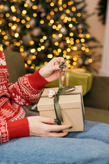 Female hands open Christmas gift box with bow on the light christmas tree background. Red sweater....