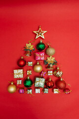 To view, the flat lay of Christmas decorations on a red background, forming a shape of a Christmas tree, Red, gold and green ball ornaments, presents, gifts, a golden bell and Christmas star