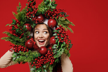 Young smiling happy cheerful caucasian woman wear Santa Claus red hat hold green Christmas wreath look aside isolated on plain red background studio portrait. Happy New Year 2022 celebration concept.