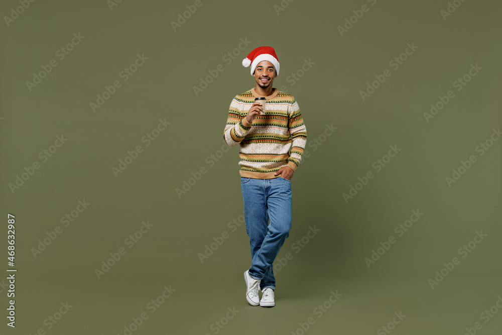 Wall mural Full body young african man in sweater red Christmas hat hold takeaway delivery craft paper brown cup coffee to go isolated on plain green khaki background. Happy New Year 2022 celebration concept