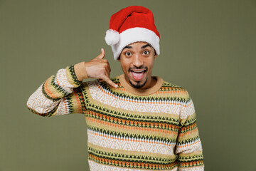 Young african man in knitted sweater red Christmas Santa Claus hat doing phone gesture like says call me back isolated on plain green khaki background studio. Happy New Year 2022 celebration concept.