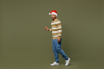 Fototapeta na wymiar Full body side view young african man 20s in knitted sweater red Christmas Santa Claus hat walking go isolated on plain green khaki background studio portrait. Happy New Year 2022 celebration concept.