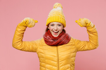 Strong sporty vivid young woman 20s years old wear yellow jacket hat mittens showing biceps muscles on hand demonstrating strength power isolated on plain pastel light pink background studio portrait