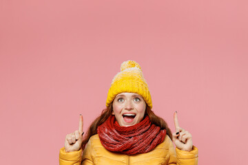 Promoter young woman 20s wears yellow jacket hat mittens pointing overhead on workspace area copy space mock up keeping mouth wide open isolated on plain pastel light pink background studio portrait.