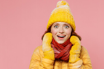 Amazed pop-eyed smiling young woman 20s years old wears yellow jacket hat mittens looking camera put hands on scarf wrapped around neck isolated on plain pastel light pink background studio portrait.