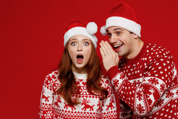 Young amazed couple friends two man woman in sweater hat whisper gossip and tell secret behind his hand share news hold face isolated on plain red background Happy New Year 2022 celebration concept