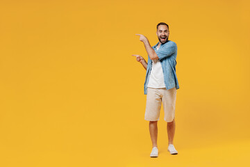 Fototapeta na wymiar Full body young smiling fun happy caucasian man 20s wearing blue shirt white t-shirt point index finger aside on workspace area mock up copy space isolated on plain yellow background studio portrait