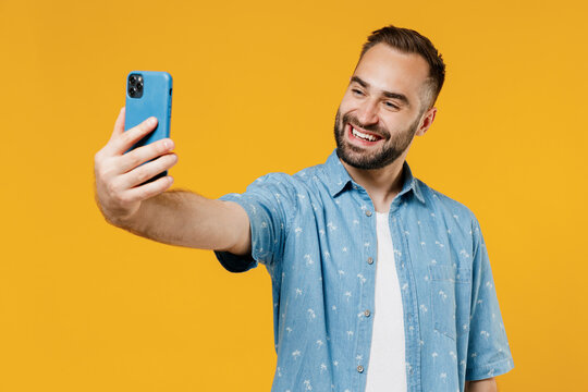 Young smiling fun caucasian friendly happy man 20s in blue shirt white t-shirt doing selfie shot on mobile cell phone post photo on social network isolated on plain yellow background studio portrait.