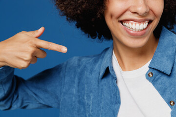 Close up cropped young smiling cheerful satisfied happy black woman in casual clothes shirt white t-shirt point index finger on smile white teeth isolated on plain dark blue color background studio