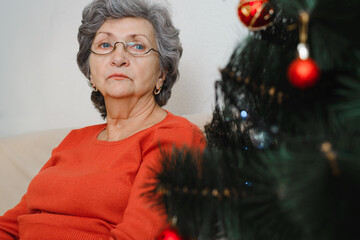 Portrait of lonely sad senior woman with glasses sitting alone at home near xmas tree and looking...
