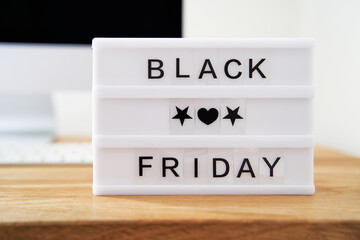 Black Friday is written on a light box. The light box is on a wooden desk. Computer in the background blurred. Black Friday sale concept.