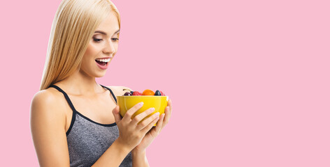 Healthy eating, vegan vegetarian raw food dieing concept image - happy excited astonished surprised shocked woman with open mouth, holding bowl plate of fruits. Blond slim girl over rose pink.