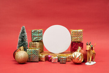 Christmas holiday red background composition with a white circle, for mock up greetings artwork. Stack of red, green and yellow presents, Christmas ornaments, reindeer and a small Christmas tree